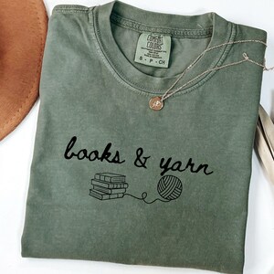 May include: An olive green t-shirt with the words "books & yarn" in black script, along with a graphic of books and a ball of yarn. A gold necklace with a small pendant rests on the shirt. A brown hat is visible in the background.