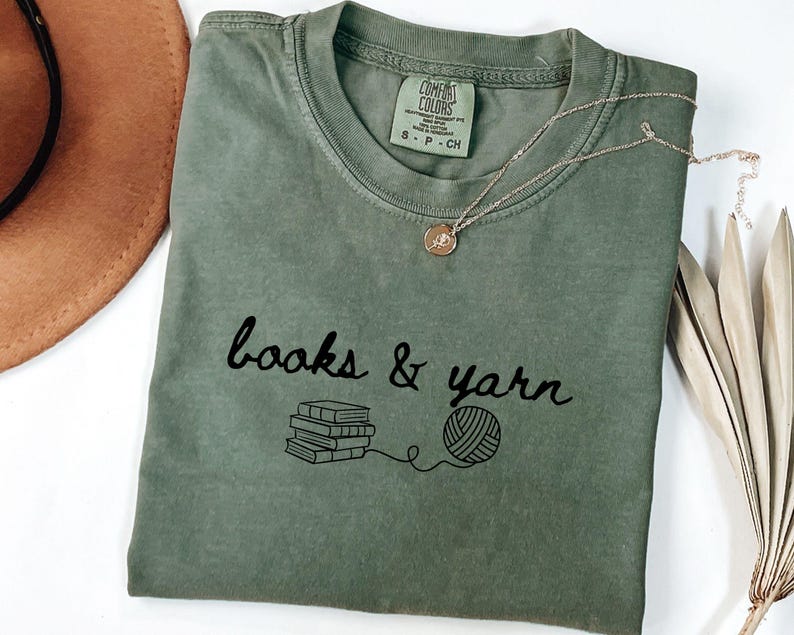 May include: An olive green t-shirt with the words "books & yarn" in black script, along with a graphic of books and a ball of yarn. A gold necklace with a small pendant rests on the shirt. A brown hat is visible in the background.