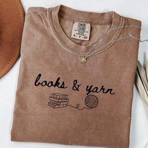 May include: A brown t-shirt with the words "books & yarn" in black script, along with illustrations of books and a ball of yarn. A silver necklace with a pendant is on the shirt. A brown hat is in the upper left corner.
