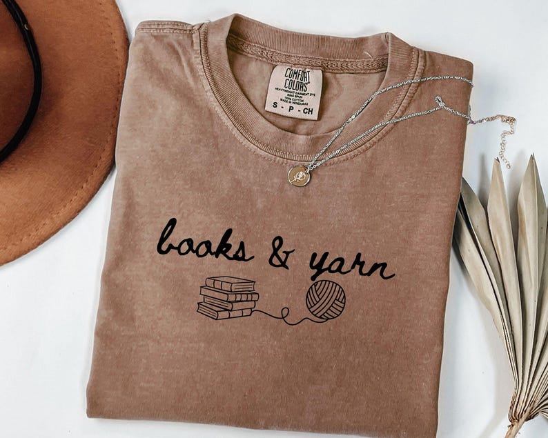 May include: A brown t-shirt with the words "books & yarn" in black script, along with illustrations of books and a ball of yarn. A silver necklace with a pendant is on the shirt. A brown hat is in the upper left corner.