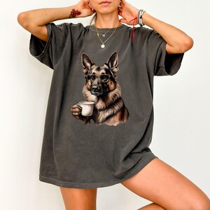 Puede incluir: Una camiseta gris oscuro de gran tama&ntilde;o con un estampado gr&aacute;fico de un perro pastor alem&aacute;n con gafas y sosteniendo una taza de caf&eacute; blanca.