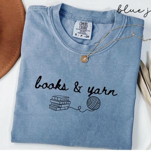 May include: A blue jean colored t-shirt with the words "books & yarn" in black script. Below the text is a stack of books and a ball of yarn connected by a line. A gold necklace with a pendant is draped on the shirt.