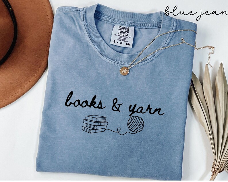 May include: A blue jean colored t-shirt with the words "books & yarn" in black script. Below the text is a stack of books and a ball of yarn connected by a line. A gold necklace with a pendant is draped on the shirt.