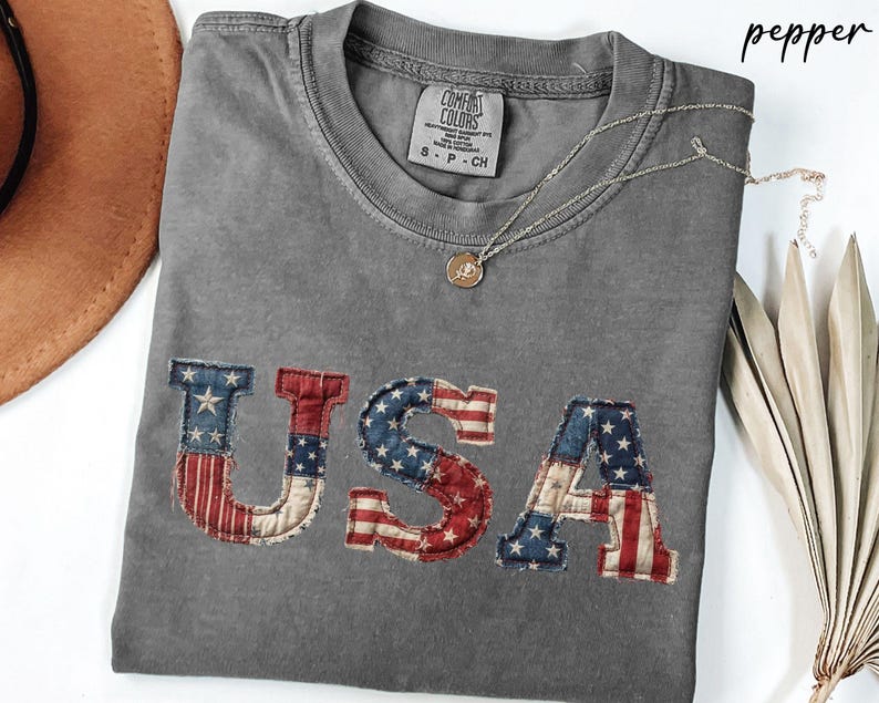May include: A gray t-shirt with the word "USA" appliqued on the front in a patriotic flag design. The shirt has a crew neck and short sleeves. A gold necklace with a pendant is draped across the shirt.