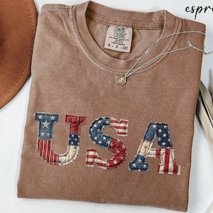 May include: Espresso-colored t-shirt with the word "USA" appliqued on the front in a patriotic flag design. The shirt has a crew neck and short sleeves. A silver necklace with a pendant is draped across the shirt.