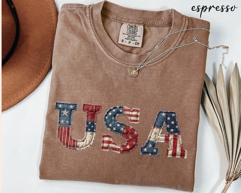 May include: Espresso-colored t-shirt with the word "USA" appliqued on the front in a patriotic flag design. The shirt has a crew neck and short sleeves. A silver necklace with a pendant is draped across the shirt.