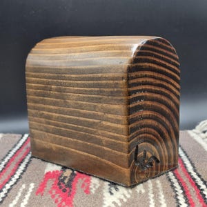 May include: A smooth, arched wooden block with a dark brown stained finish, showcasing prominent natural wood grain patterns. The piece features a curved top and a flat base, with a small, dark, hand-carved signature mark visible on the lower right side.