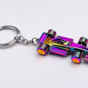 Könnte beinhalten: Ein schillernder Schlüsselanhänger in Form eines Formel-1-Rennwagens. Der Miniaturwagen mit vier Rädern und aerodynamischem Design ist an einem silbernen Schlüsselring befestigt. Die metallische Oberfläche des Autos zeigt ein Spektrum an Farben, darunter Lila, Blau und Gold.