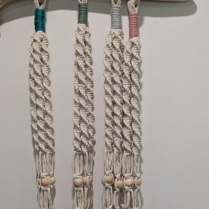 May include: Four macrame plant hangers in a row, each with a different colored top: teal, green, gray, and pink. The off-white rope is knotted in a spiral pattern, with wooden beads and wooden rings at the top.