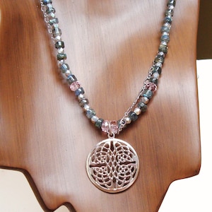 Peut inclure: Un collier pendentif argenté avec un motif ajouré circulaire. Le collier présente une chaîne de perles avec des perles grises, vertes et roses. Le pendentif est suspendu à une chaîne délicate, exposé sur un buste en bois.
