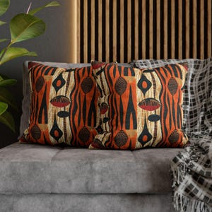 May include: Two decorative throw pillows with an abstract geometric pattern in shades of orange, black, and white. The pillows are on a gray couch with a black and white patterned throw blanket.
