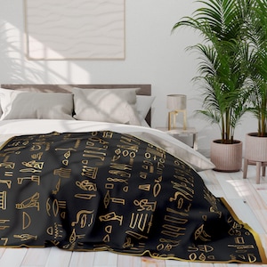 Pode incluir: Cobertor preto e dourado com um padrão de hieróglifos egípcios. O cobertor está dobrado em uma cama com roupa de cama branca e uma cabeceira de madeira.