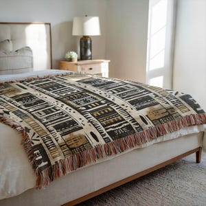 May include: A woven throw blanket with a geometric pattern in black, tan, and brown, fringed at the bottom. The blanket is draped over a bed with a neutral-colored headboard and a wooden frame.