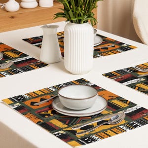 May include: A table setting with patterned placemats in orange, yellow, black, and red geometric designs. The setting includes white bowls, plates, and silverware, alongside a white vase with green foliage. The table is covered with a white tablecloth.