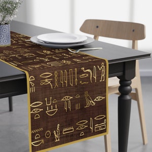 May include: A dark brown table runner featuring a repeating pattern of gold-coloured Egyptian hieroglyphs. The fabric has a textured appearance and is finished with a solid gold-coloured border along the edges of the rectangular textile piece.