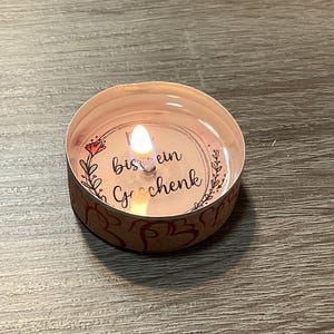 May include: A small pink tea light candle with a floral design and the text "Bis ein Geschenk" in black lettering.