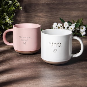 Puede incluir: Dos tazas de cerámica. Una es rosa con el texto "We love you, Boppy!" y un corazón. La otra es blanca con el texto "MAMMA" y un corazón. Ambas tazas tienen una base marrón y un asa curva.