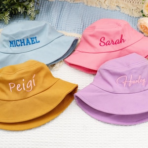May include: Four brightly coloured bucket hats, each embroidered with a name. The hats are in shades of blue, pink, yellow, and purple. The names embroidered are Michael, Sarah, Peigí, and Harley. The hats are on a white surface with flowers.