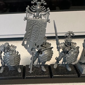 May include: A set of six unpainted plastic miniature figures of skeletal warriors, including a king, a standard bearer, and four soldiers. The figures are posed in a variety of combat stances, holding swords and shields.