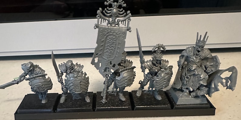 May include: A set of six unpainted plastic miniature figures of skeletal warriors, including a king, a standard bearer, and four soldiers. The figures are posed in a variety of combat stances, holding swords and shields.