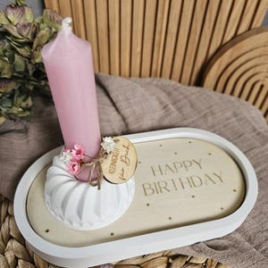 Può includere: Un vassoio ovale in legno bianco con bordo bianco, che contiene una candela rosa e un portacandele bianco scanalato. Il vassoio ha incise le parole "HAPPY BIRTHDAY", insieme a un piccolo cartello di legno con "Kleinigkeiten für Dich".