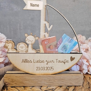 Puede incluir: Caja de madera para dinero en forma de velero con una jirafa, un león y un pequeño tigre en la parte delantera. La caja tiene una ranura para el dinero y un letrero que dice "Nombre" en la parte superior. La caja también tiene el texto "Alles Liebe zur Taufe 23.03.2025" en la parte inferior.