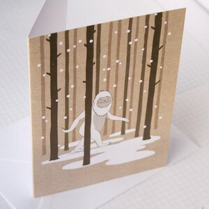 Puede incluir: Una tarjeta marr&oacute;n con un Yeti blanco caminando por un bosque nevado. El Yeti sonr&iacute;e y tiene una barba blanca. El bosque est&aacute; formado por &aacute;rboles marrones con nieve blanca cayendo.
