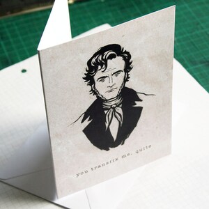 May include: A black and white illustration of a man with a serious expression. He is wearing a dark suit and a white cravat. The text "you transfix me, quite" is printed below the illustration.