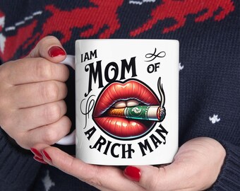 May include: White ceramic mug with the text "I AM MOM OF A RICH MAN". The mug features a graphic of red lips holding a lit cigarette with a dollar bill inside. The mug is held by a person.