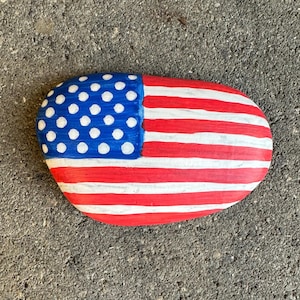 May include: A hand-painted, oval-shaped rock depicting the American flag. The design includes a blue section with white dots and red and white stripes. Ideal for patriotic decor or as a unique gift.