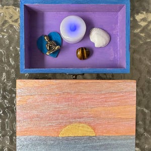 Puede incluir: Una caja de madera rectangular con exterior azul e interior morado. La tapa presenta una pintura de un atardecer con un sol amarillo sobre un horizonte gris. Dentro hay una pequeña figura de Buda dorada, un corazón azul, una concha blanca y una piedra ojo de tigre.