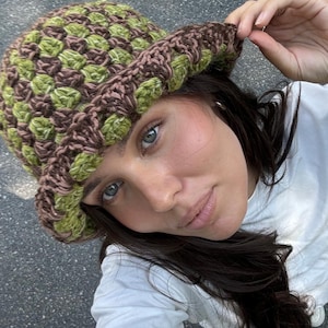 May include: A crochet bucket hat in green and brown. The hat has a textured stitch and a wide brim. This handmade hat is a fashionable accessory for sun protection, adding a unique touch to any outfit. The hat is a stylish accessory.