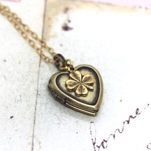 May include: A gold-colored necklace with a heart-shaped locket pendant. The locket features a four-leaf clover design. The necklace is on a vintage-style background with handwritten text.
