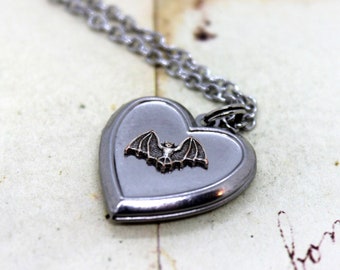 Può includere: Una collana con un ciondolo a forma di cuore argentato con un piccolo ciondolo a forma di pipistrello. Il ciondolo è aperto e l'interno è vuoto.