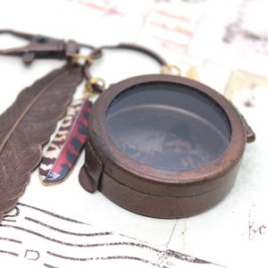 May include: A bronze-colored round locket with a clear glass window, attached to a keychain with a lobster clasp. The locket is adorned with a large feather and smaller decorative feathers in red, black, and white.