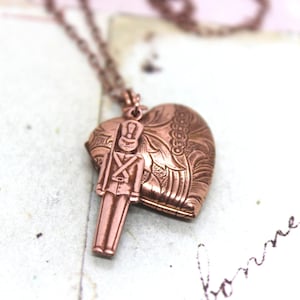 May include: A copper-toned heart-shaped locket necklace with a detailed floral design. The locket features a small copper-toned toy soldier charm.