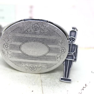 May include: A silver-toned locket with an oval center and floral accents. A small, dark-colored soldier charm stands beside the locket. The locket has a striped design and is on a light-colored surface.