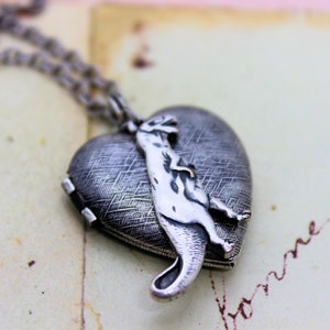 May include: A silver heart-shaped locket necklace with a detailed animal figure attached to the front. The locket has a textured surface and a hinged opening.