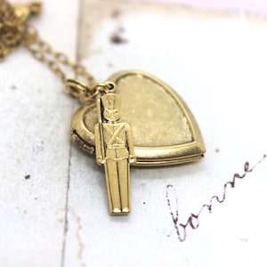 May include: A gold heart-shaped locket necklace with a small gold toy soldier charm. The locket has a hinged opening and is attached to a gold chain.