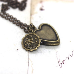 May include: An antique bronze-coloured necklace with a heart-shaped locket and a round charm bearing the Scorpio zodiac symbol. The necklace has a fine chain, set against a vintage paper background.