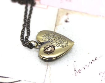 May include: An antique gold heart-shaped locket necklace. The locket has floral and ladybug detailing. The chain is dark bronze. The locket is closed and rests on a white surface with faint script.