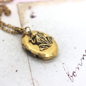 Könnte beinhalten: Goldene Halskette mit einem Anhänger in Form eines Frosches. Das Medaillon ist oval und hat einen Klappdeckel. Der Frosch sitzt auf dem Medaillon.
