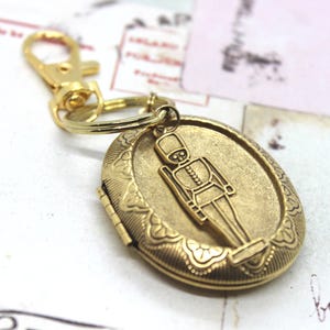 May include: A gold-colored metal keychain featuring an oval locket with a soldier design. The locket is engraved with floral patterns and opens to reveal a space for a small photo or keepsake. The keychain has a lobster clasp.