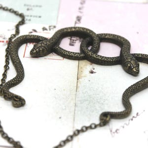 May include: A bronze-toned snake necklace with three intertwined snake pendants. The necklace features a delicate chain and a small clasp. The snakes have detailed scales and are positioned on a vintage paper background.