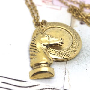 May include: A gold-colored pendant necklace featuring a horse head chess piece and a round locket. The horse head is detailed with a mane and facial features. The locket has a floral design around the edge. The necklace is on a gold chain.