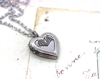 Può includere: Collana con ciondolo a forma di cuore in argento con un design a forma di pipistrello sul davanti. Il ciondolo è aperto e l'interno è visibile.