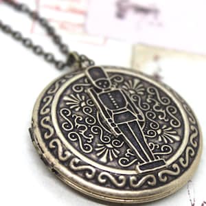 May include: A silver-coloured locket necklace with an intricate design. The round pendant features a soldier figure in the centre, surrounded by an ornate pattern. The necklace has a chain and clasp.