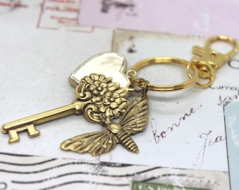 Può includere: Un portachiavi color oro con un ciondolo a forma di cuore, chiave, fiore e farfalla. La chiave ha un design floreale dettagliato e la farfalla ha motivi alari intricati. Il portachiavi include un fermaglio e un anello per attaccare le chiavi.