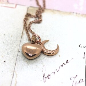 May include: Rose gold necklace featuring a heart and crescent moon pendant. The heart is smooth and polished, while the moon has a textured surface. The chain and pendants are a warm, metallic color, creating a romantic aesthetic.