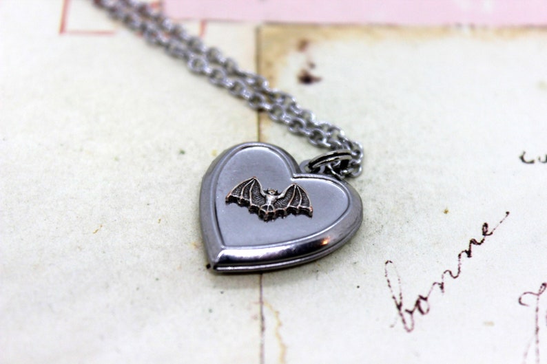 May include: A silver heart-shaped locket necklace with a small bat charm. The locket is open and the inside is blank.